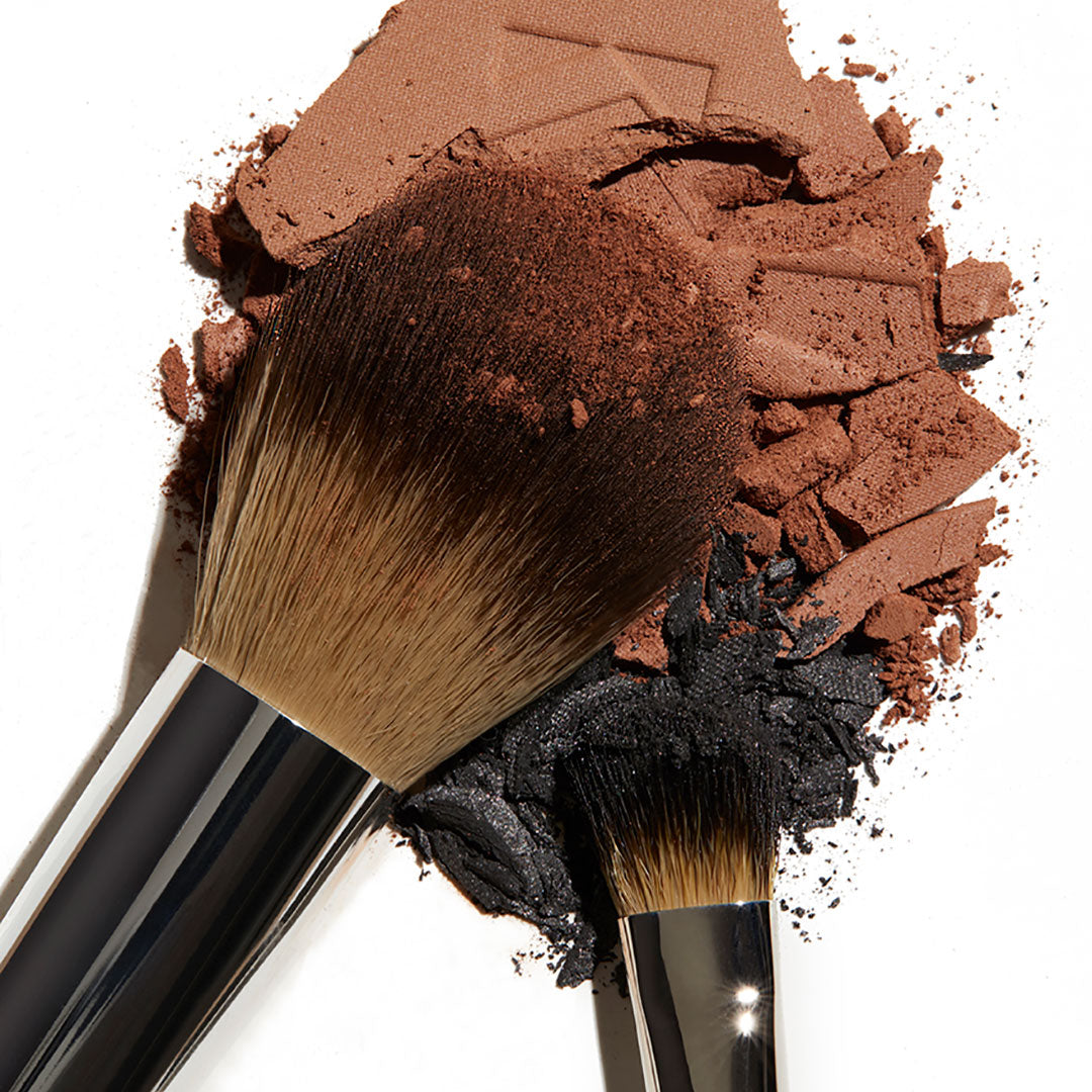 Brown and black makeup crumbled on a white background with two makeup brushes