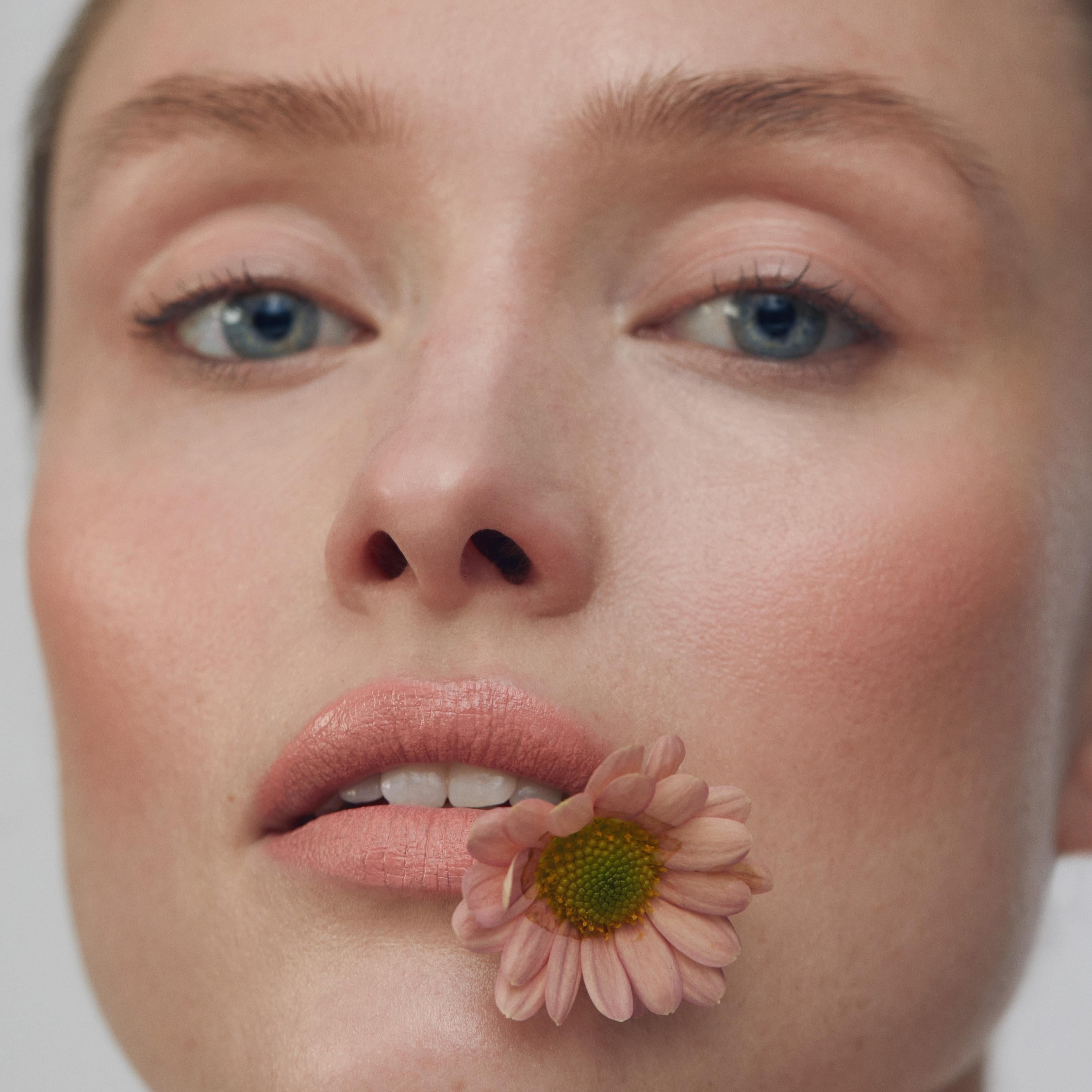 Close up of a person’s face with a fair skin tone, wearing cream blush and holding a light pink flower in their mouth