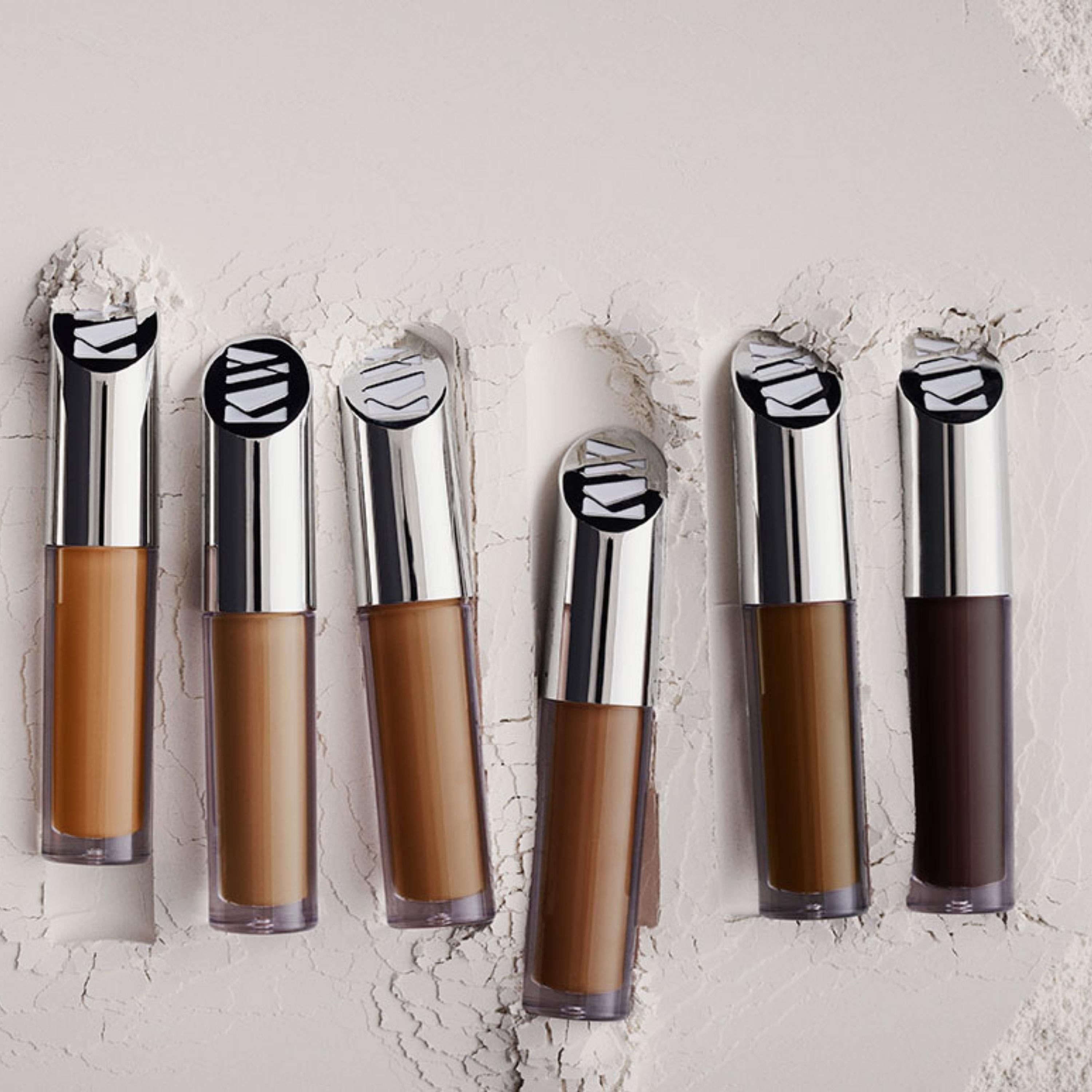 Set of six makeup concealer bottles with silver caps on a textured light background