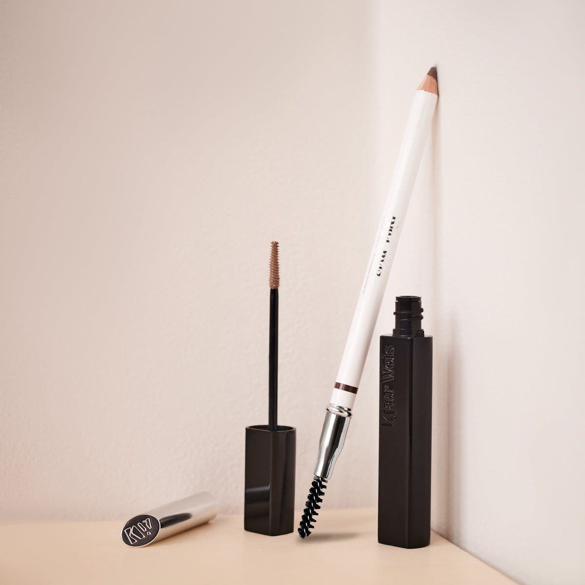 Makeup products, including an eyebrow pencil and brow gel, on a beige surface with a light background.
