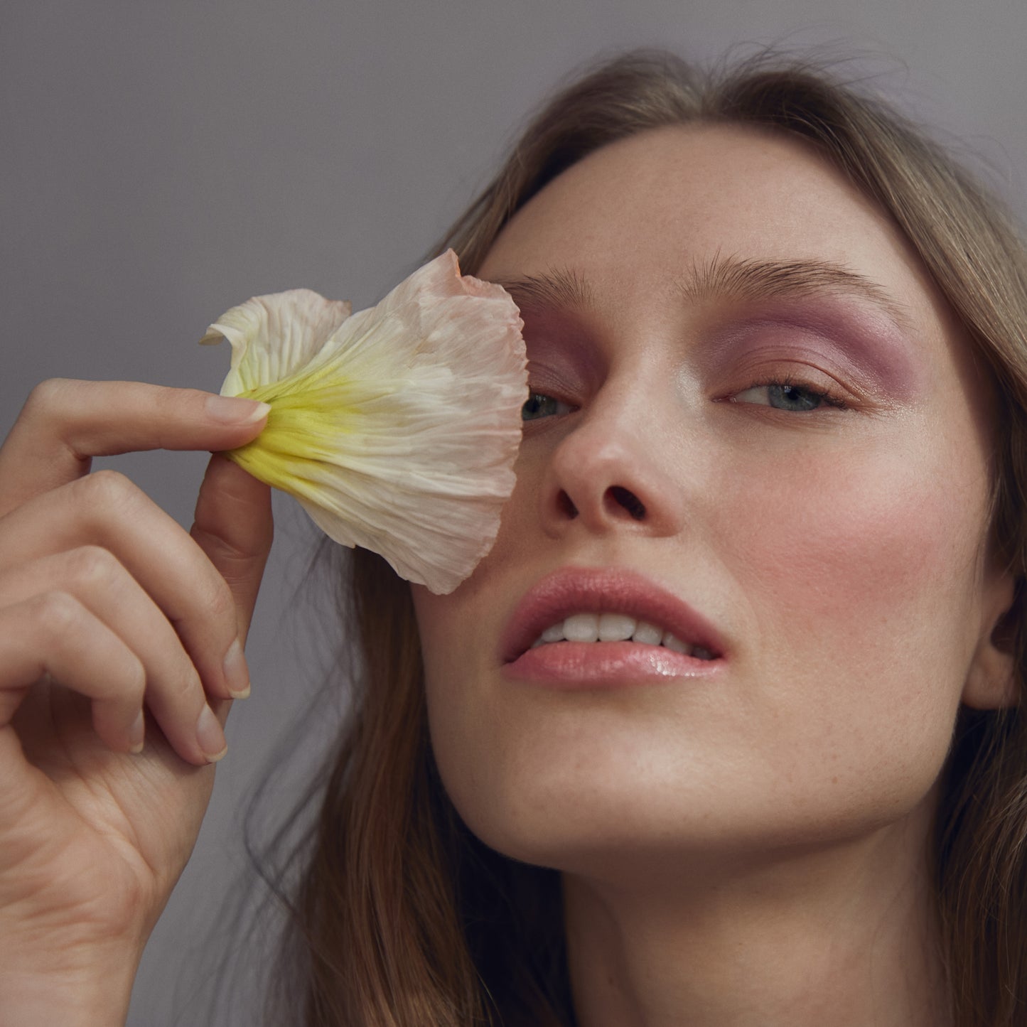 Close up of a person’s face with a fair skin tone wearing cream blush and holding a white and pink flower