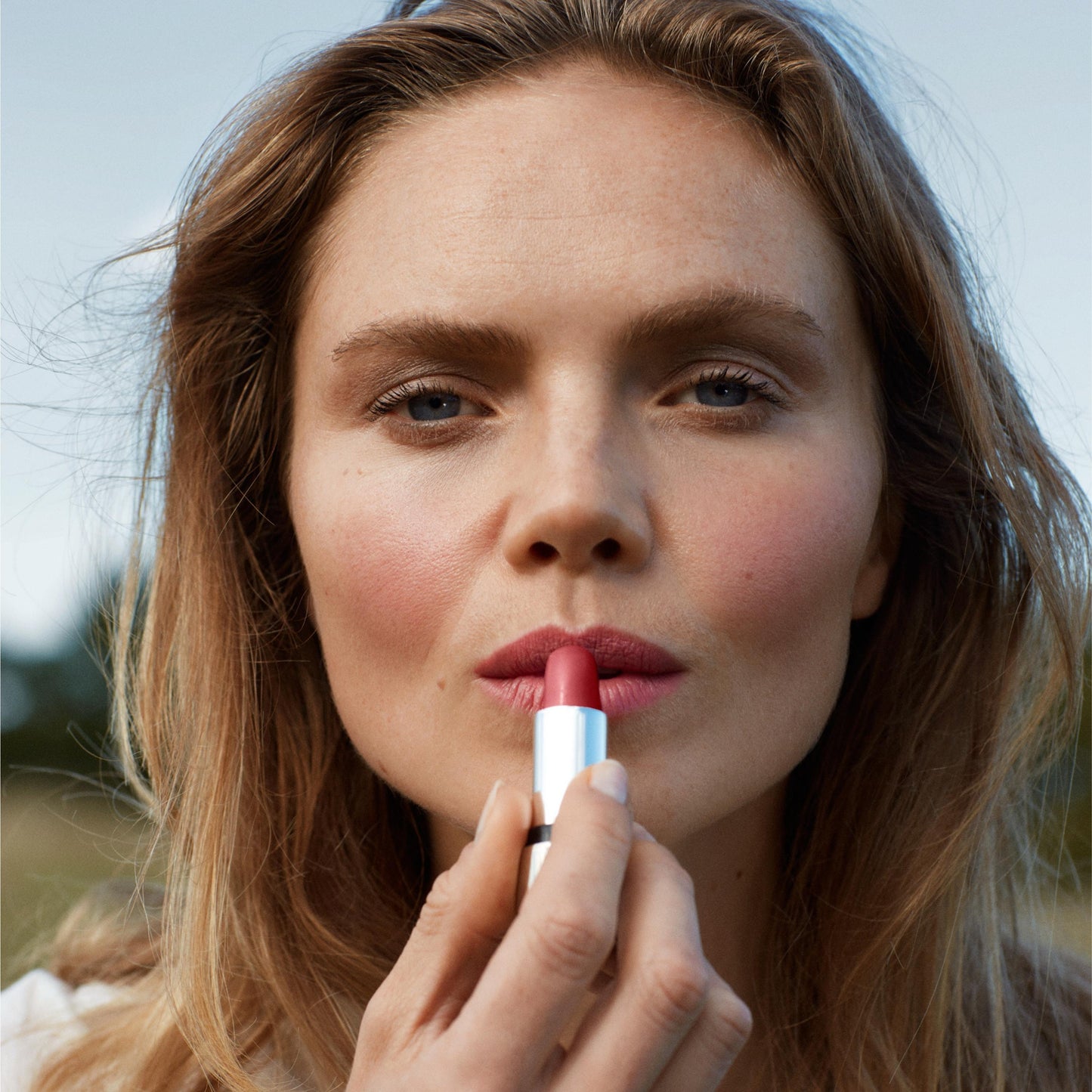 Close up of a person’s face with a fair skin tone wearing cream blush and applying pink lipstick