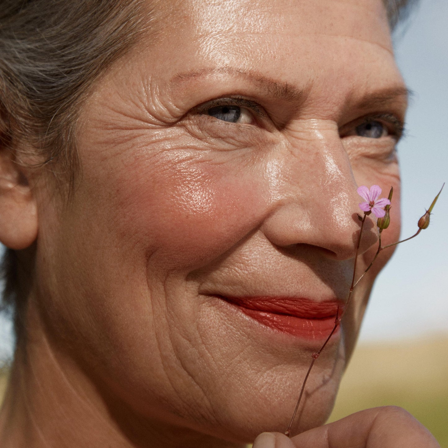 Close up of a person’s face with a fair skin tone, wearing cream blush and holding a pink flower