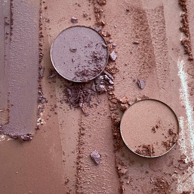 A lilac circle of eye shadow and a beige circle of eye shadow on a background covered in lilac and beige eye shadow