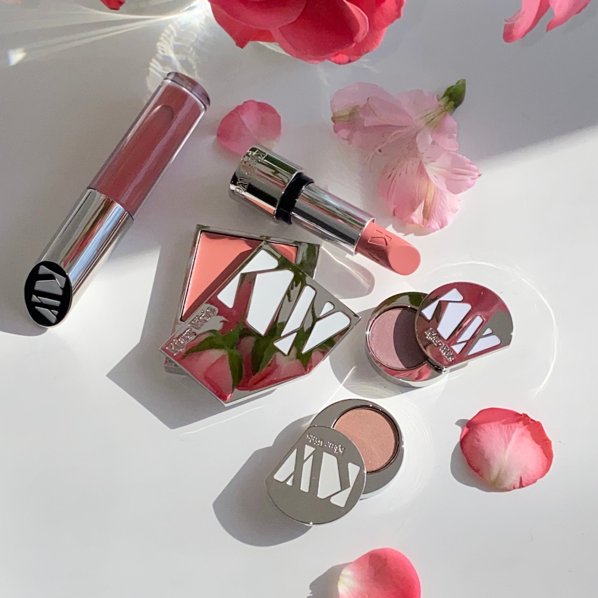 Two KW eye shadow palettes alongside KW lipstick, lip gloss, and cream glow, surrounded by pink flower petals 