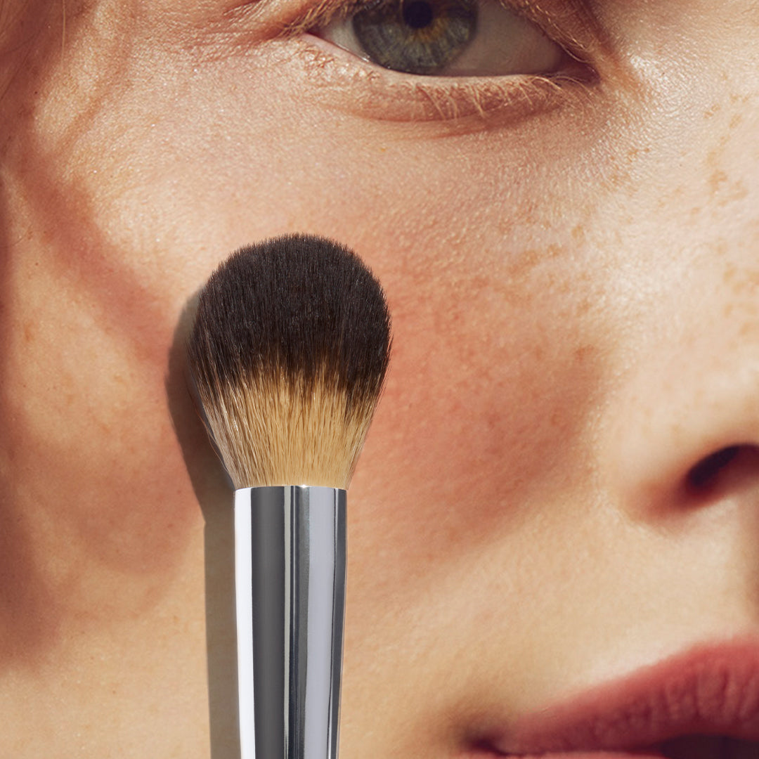 close up of a person’s face with a large rounded makeup brush on the cheek