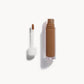 An open bottle of concealer on its side next to a white refill wand
