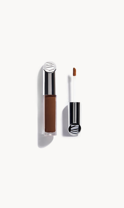Kjaer Weis concealer in a clear bottle with silver metal top next to the concealer wand