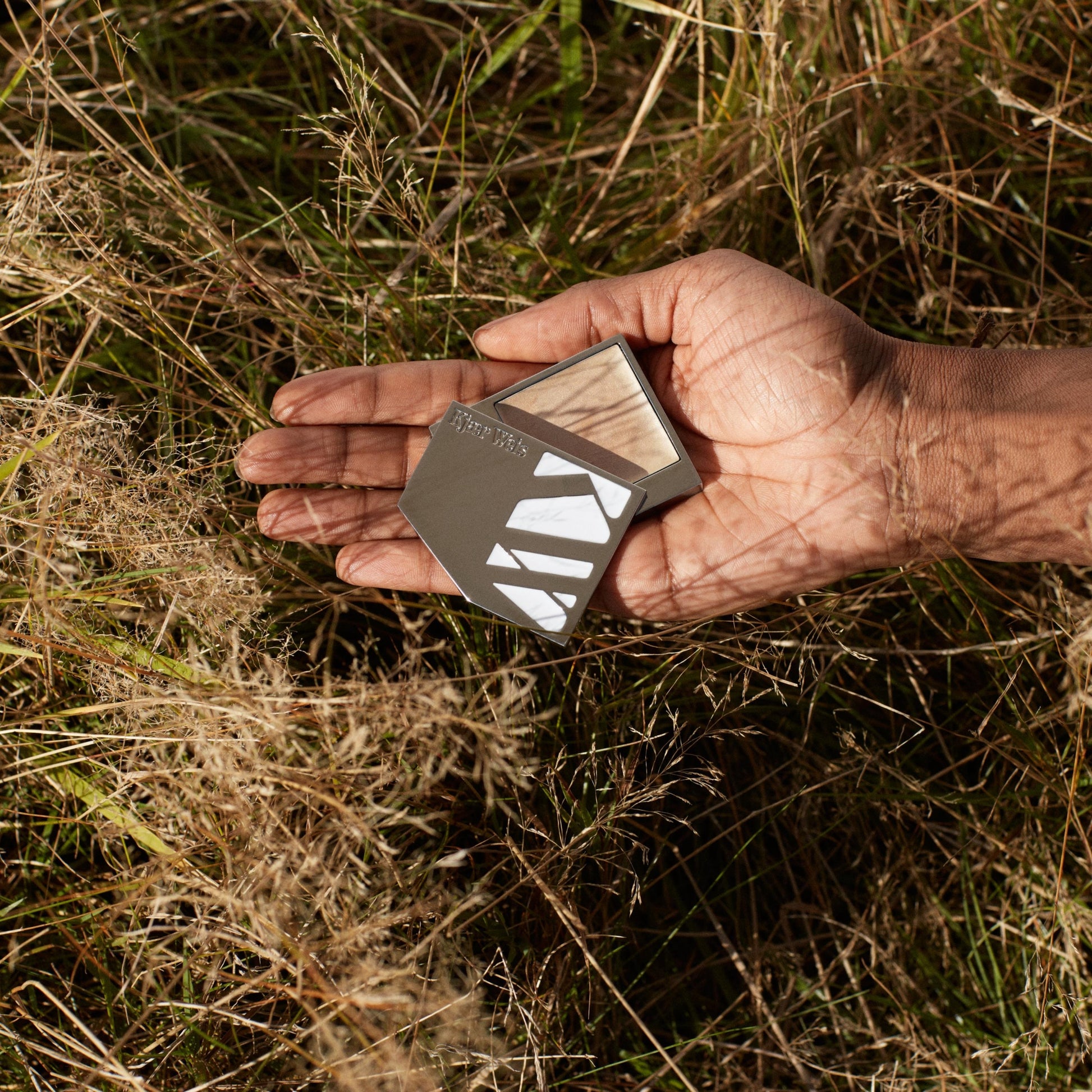 close up of a hand holding a silver palette of cream highlighter on grass